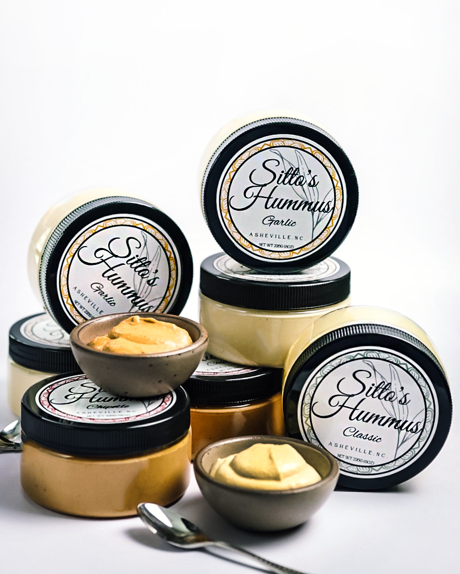 Containers of Sitto's Hummus, garlic flavor, in glass jars with black lids, along with small bowls of hummus, and spoons on a white background.