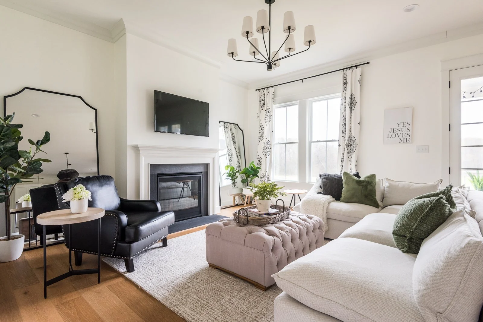 Bright living room with white walls, large windows, a white sectional sofa with green and black pillows, a tufted pink ottoman, a black leather armchair, a TV above a fireplace, and potted plants.