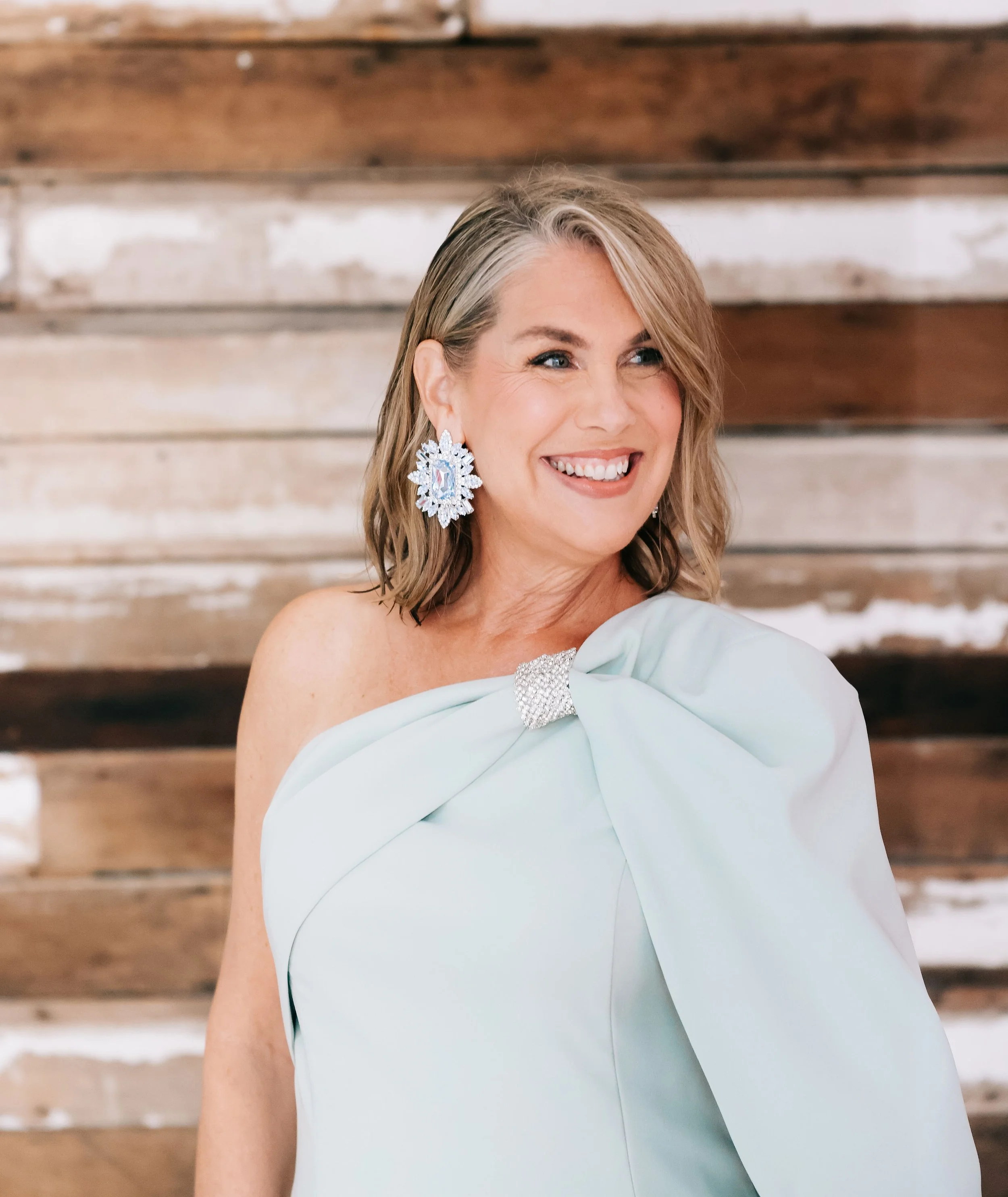 A woman with grayish-brown hair and blue eyes, wearing large snowflake-shaped earrings and a light blue off-shoulder dress, smiling against a rustic wooden background.