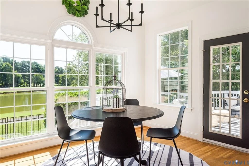 A bright dining area with large windows overlooking a pond and outdoor furniture. A black round table with four black chairs, a chandelier, and a striped rug, with a door leading outside.
