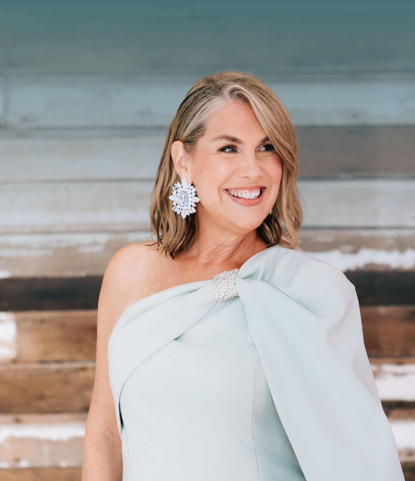 Broker/Owner Jan Copeland with shoulder-length blonde hair, wearing a seafoam green off-shoulder dress and large snowflake-shaped earrings, smiling, standing in front of a rustic wooden wall.