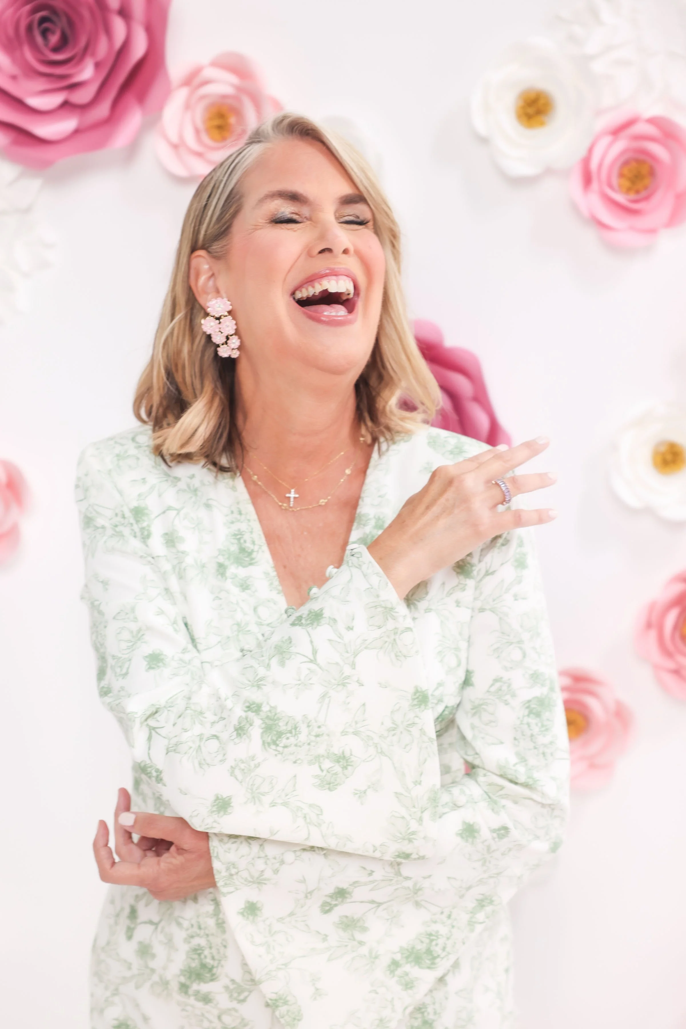 A woman with blonde hair wearing pink floral earrings and a pearl cross necklace, smiling and laughing with her eyes closed, wearing a white dress with green floral patterns, in front of a backdrop decorated with large pink and white paper flowers.