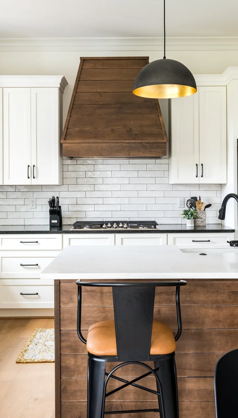 Modern kitchen with white cabinets, white subway tile backsplash, and black countertop. There is a black and gold pendant light hanging from the ceiling, a wooden range hood, and a kitchen island with a white countertop and a wooden front. A black chair with an orange cushion is at the island, and small appliances and utensils are visible on the counter.
