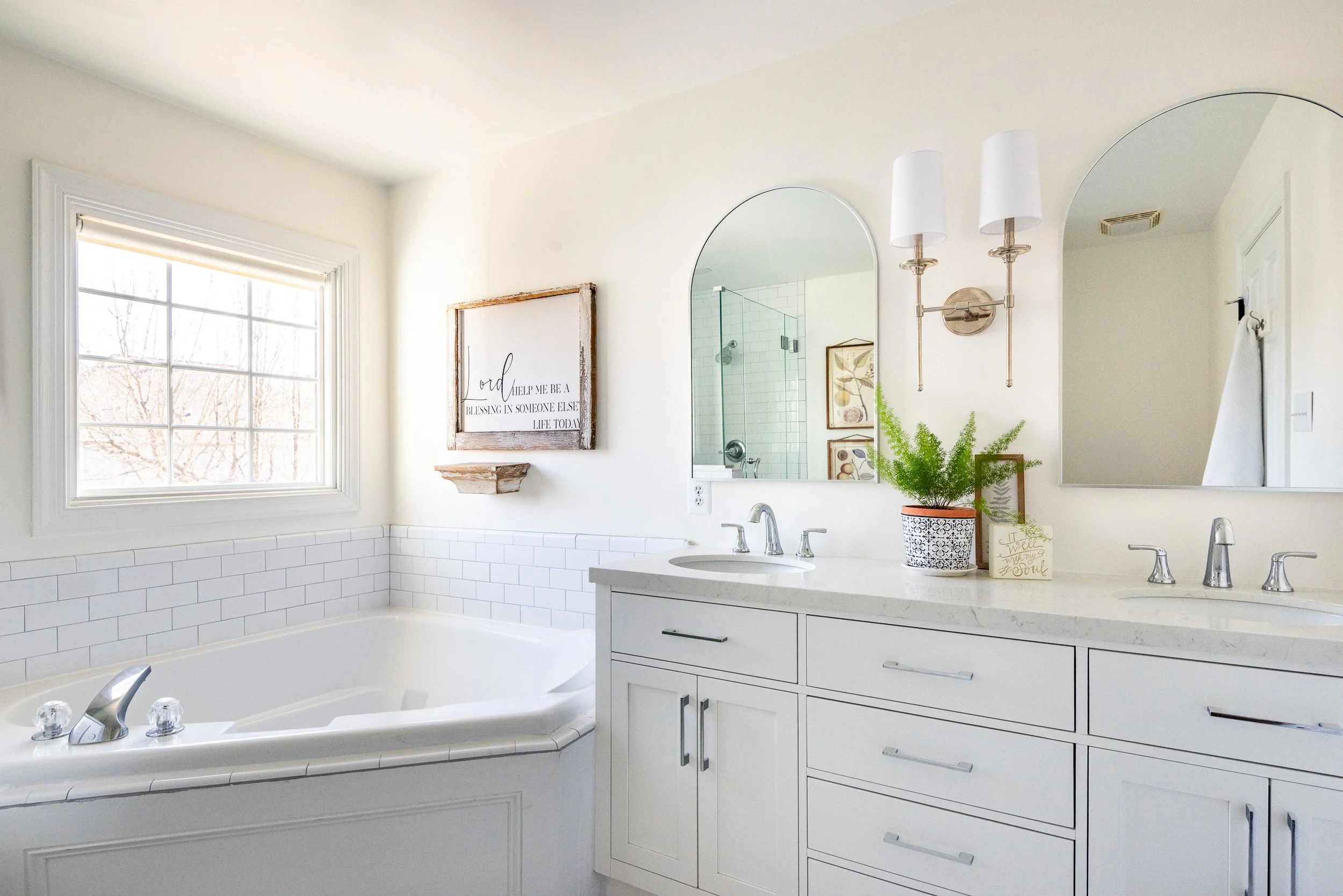 Bright bathroom with a large mirror, two wall-mounted light fixtures, a window, a white countertop with two sinks, a potted plant, framed artwork, and a shower with glass door in the background.