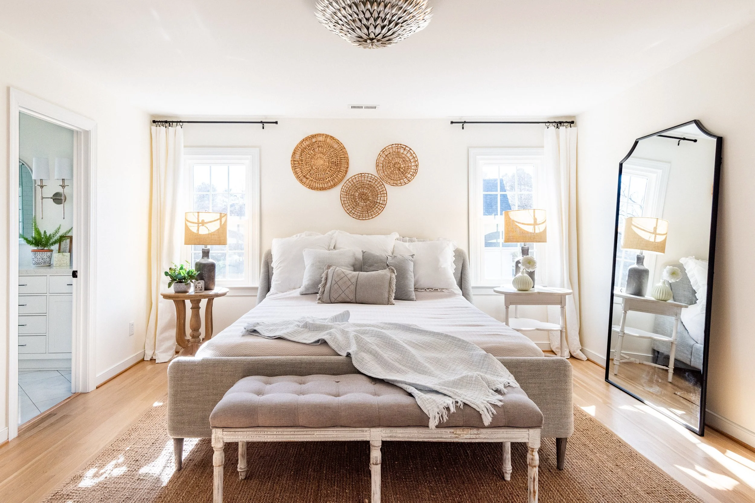 Bright bedroom with white walls, a bed with white bedding, two bedside tables with lamps, a large mirror, and decorative wall baskets.