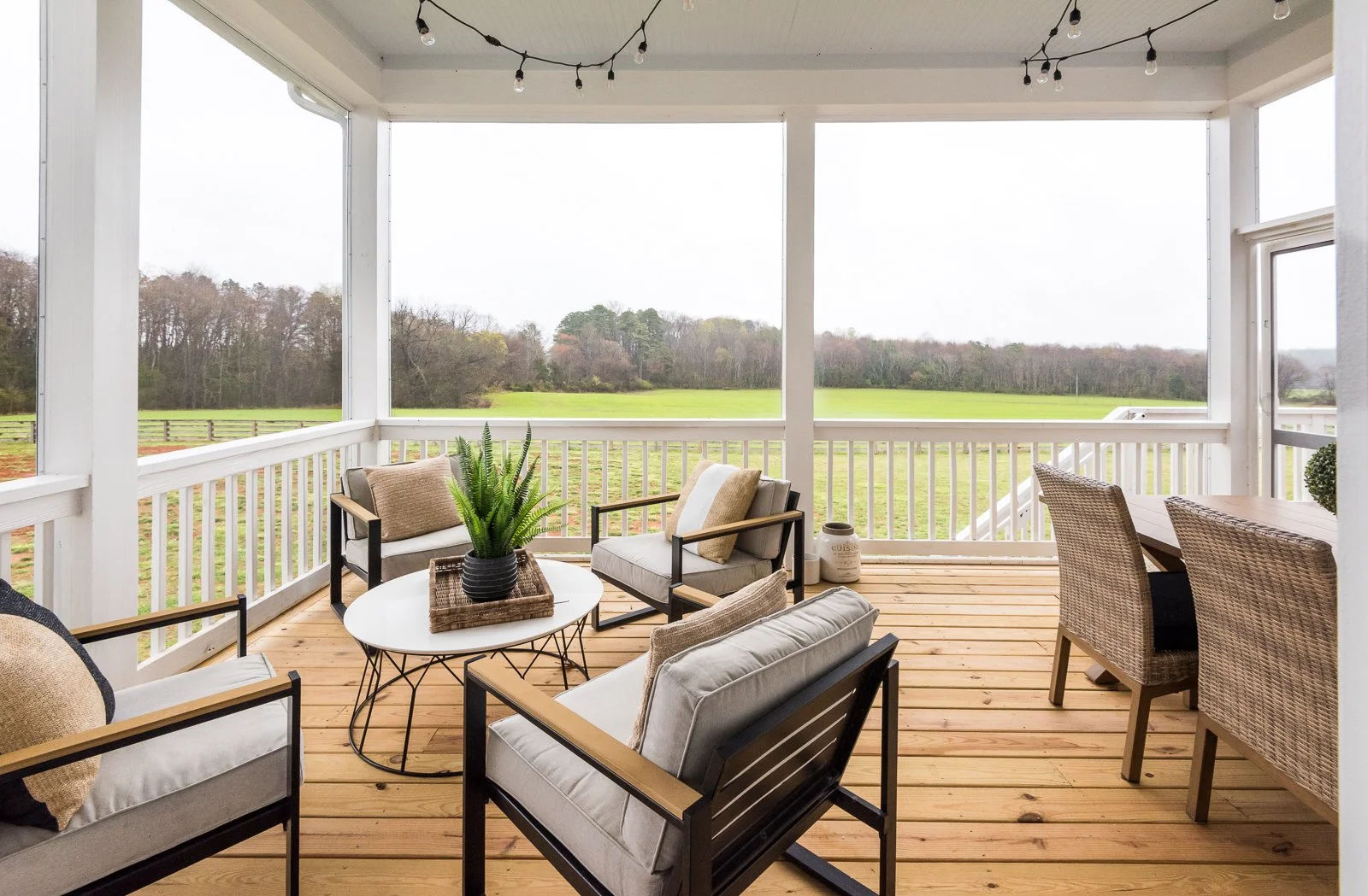 Porch with beige and white chairs, a white round table with a potted plant, and string lights, overlooking a green field and trees in the distance.