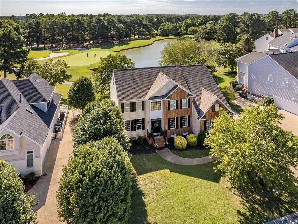 Aerial view of a modern house with a brown brick and white exterior, surrounded by green trees, with a curved walkway leading to the front door, located in a suburban neighborhood near a golf course and water feature.