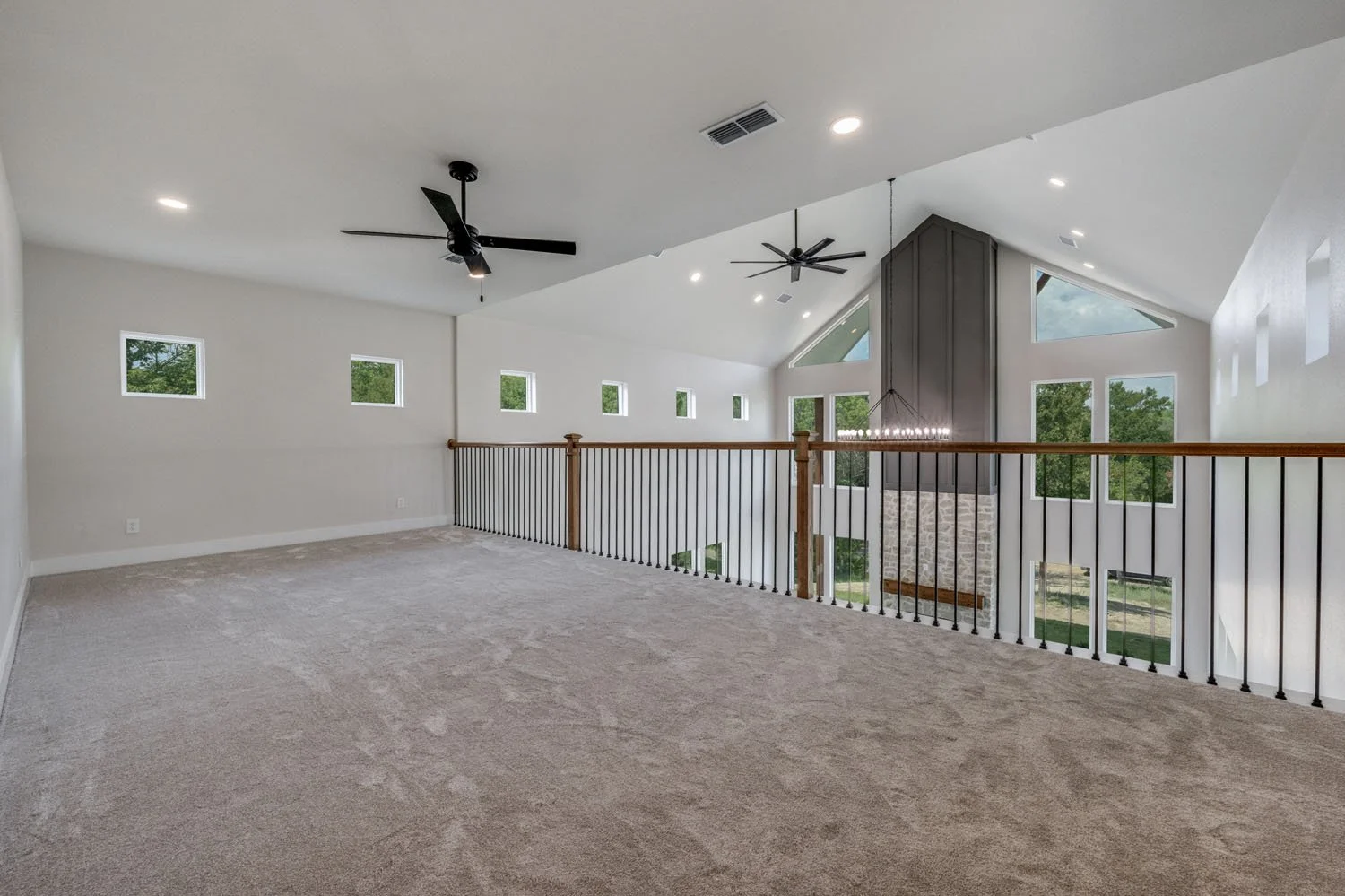 A carpeted loft area overlooking a living space, featuring a dark metal railing with wood accents, two ceiling fans, and a high vaulted ceiling.