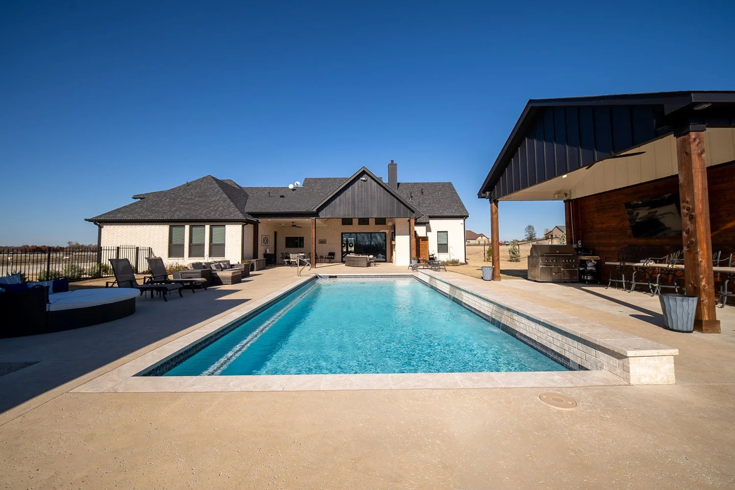 A backyard view of a custom home featuring a rectangular swimming pool, concrete patio, and a covered outdoor living area with a built-in grill.