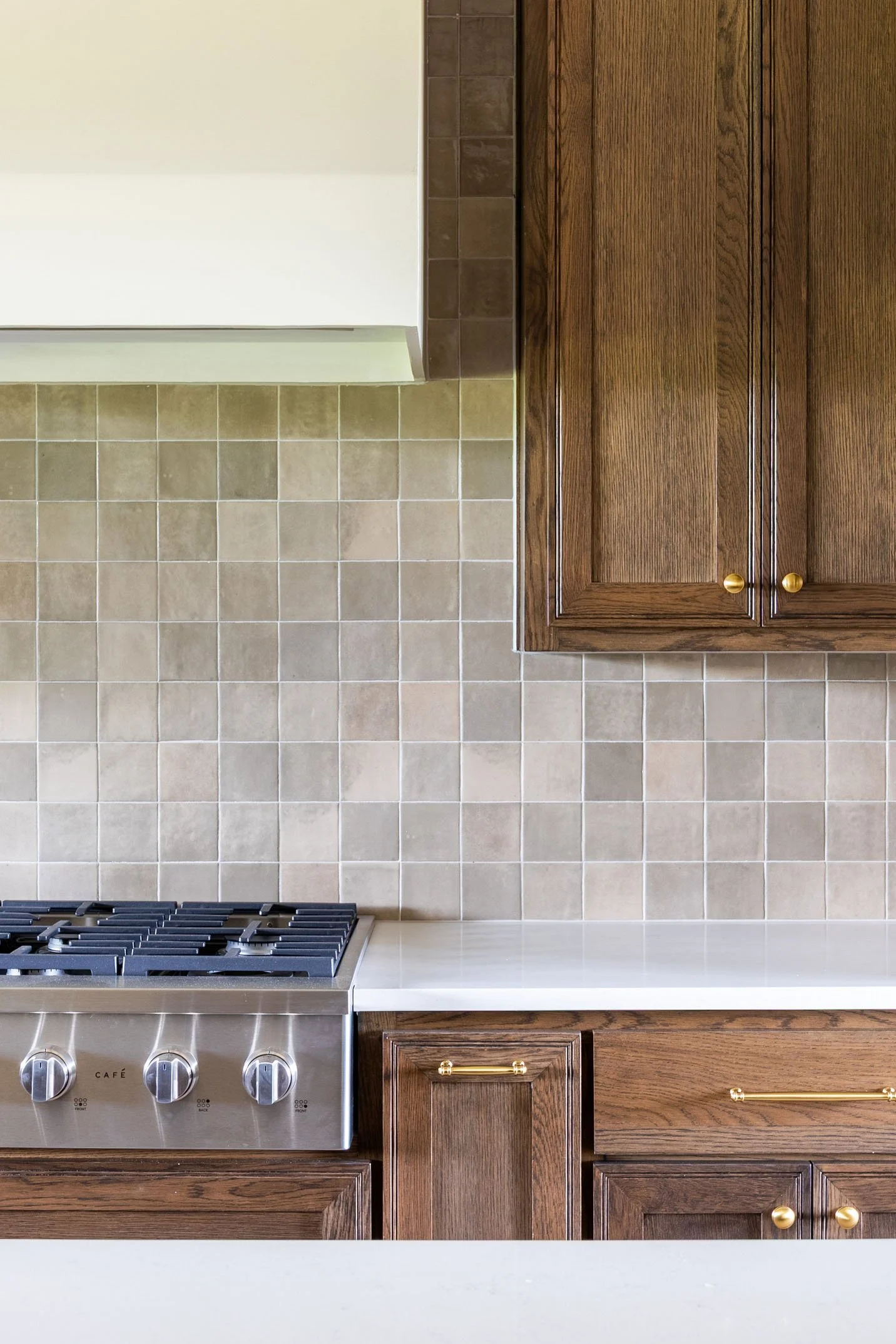 A close-up of a kitchen area featuring a stainless steel gas range, white quartz countertops, a square-tiled neutral backsplash, and dark wood cabinets with brass hardware.