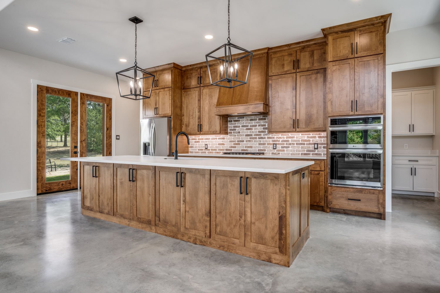 A modern kitchen featuring rustic stained wood cabinetry, a large kitchen island with white countertops, and a brick tile backsplash.