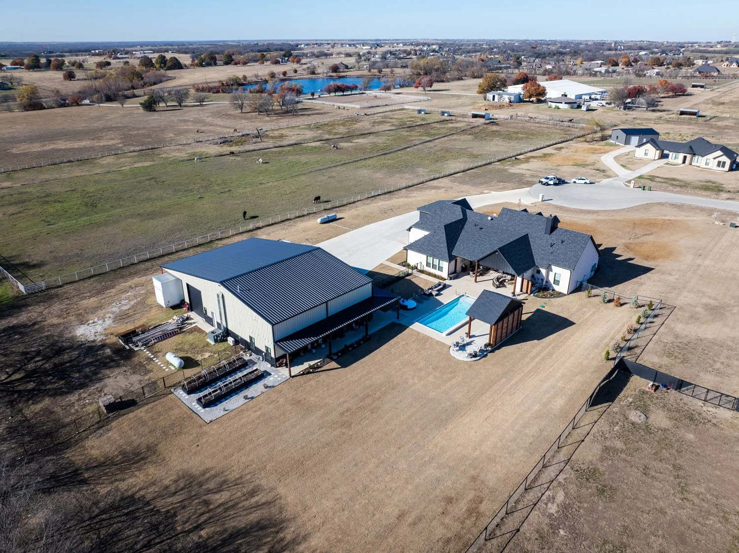 An aerial view of a rural property featuring a custom home with a black roof, a swimming pool with a covered patio area, and a large metal workshop.