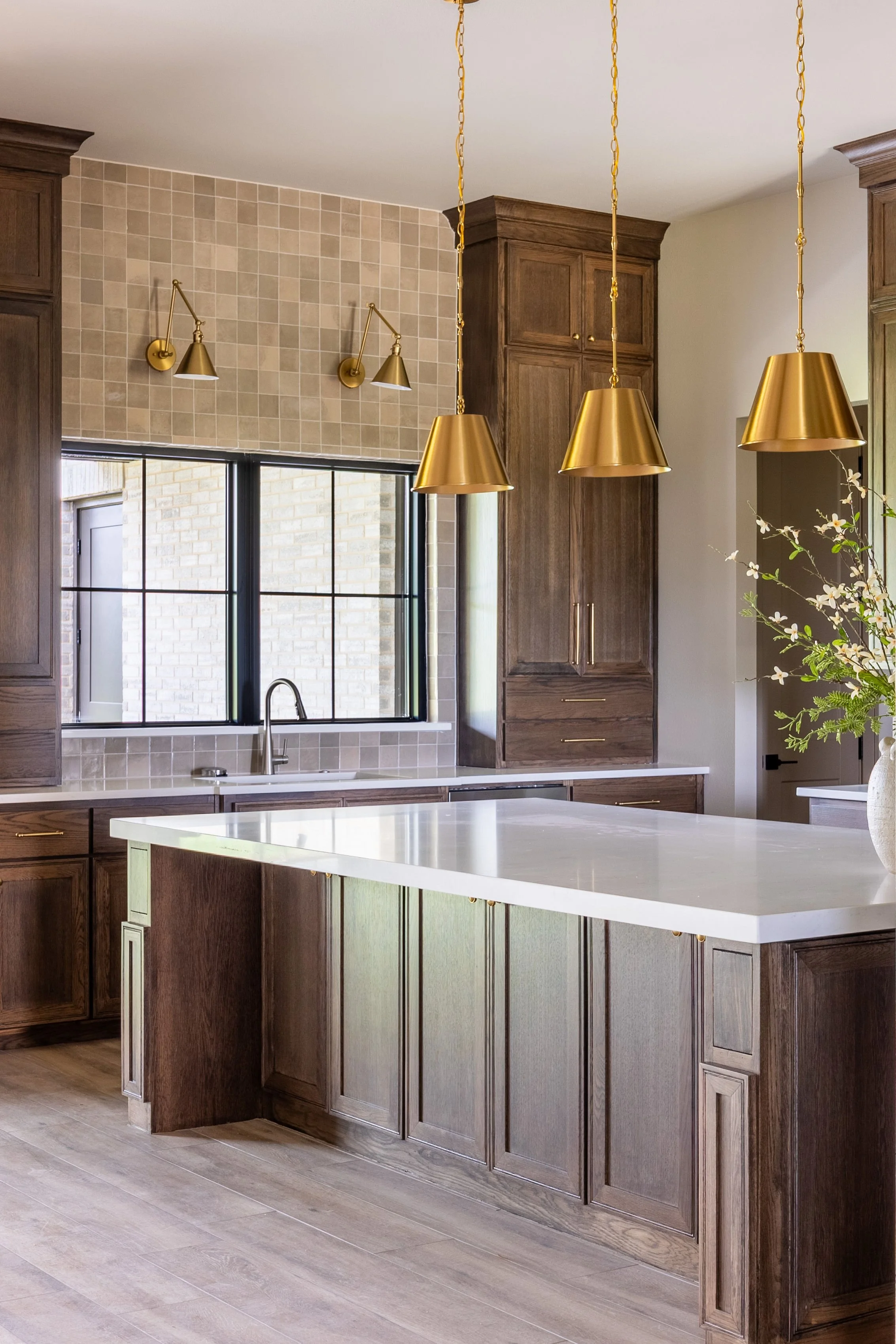 Modern kitchen with dark wood cabinets, beige tiled wall, large kitchen island with white countertop, gold pendant lights, and a window over the sink.