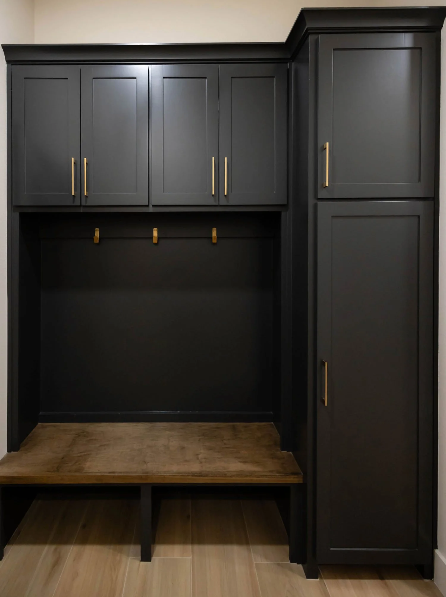 A mudroom built-in unit featuring dark charcoal cabinetry, a wood bench seat, brass hardware, and wall hooks.
