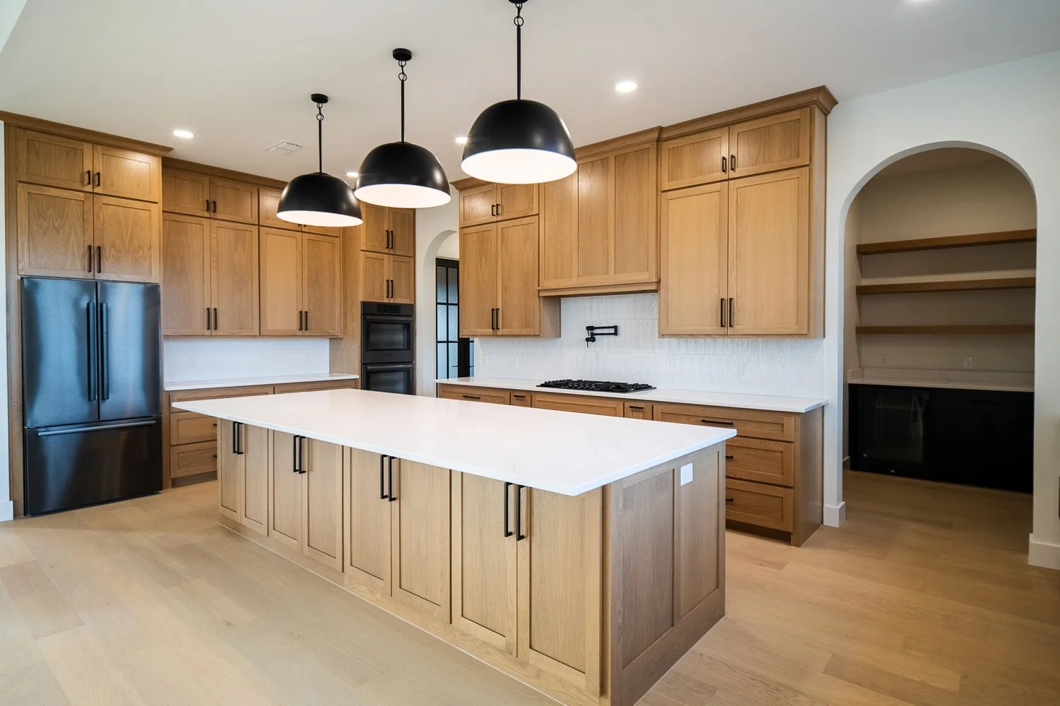 A spacious, modern kitchen featuring natural oak cabinetry, a large central island with a white quartz countertop, black pendant lighting, and an arched entryway leading to a pantry with open shelving.