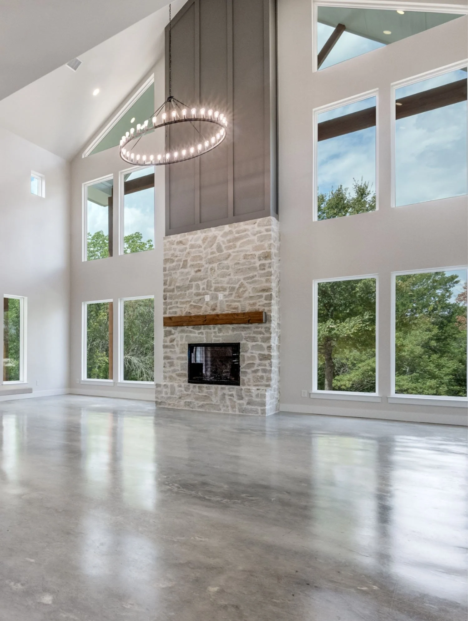 A spacious living room with polished concrete floors, a double-height vaulted ceiling, a large stone fireplace, and floor-to-ceiling windows