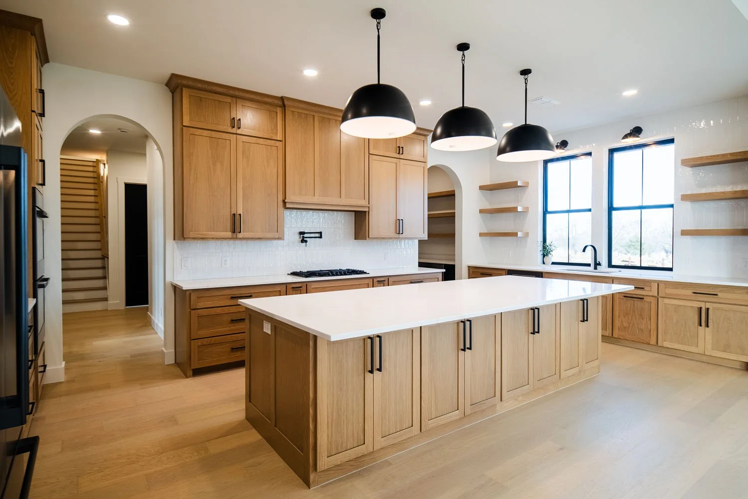 An open-concept kitchen and living area showcasing warm oak cabinets, a wide white quartz kitchen island, sleek black pendant lights, and arched entryways highlighting architectural detail and custom shelving.