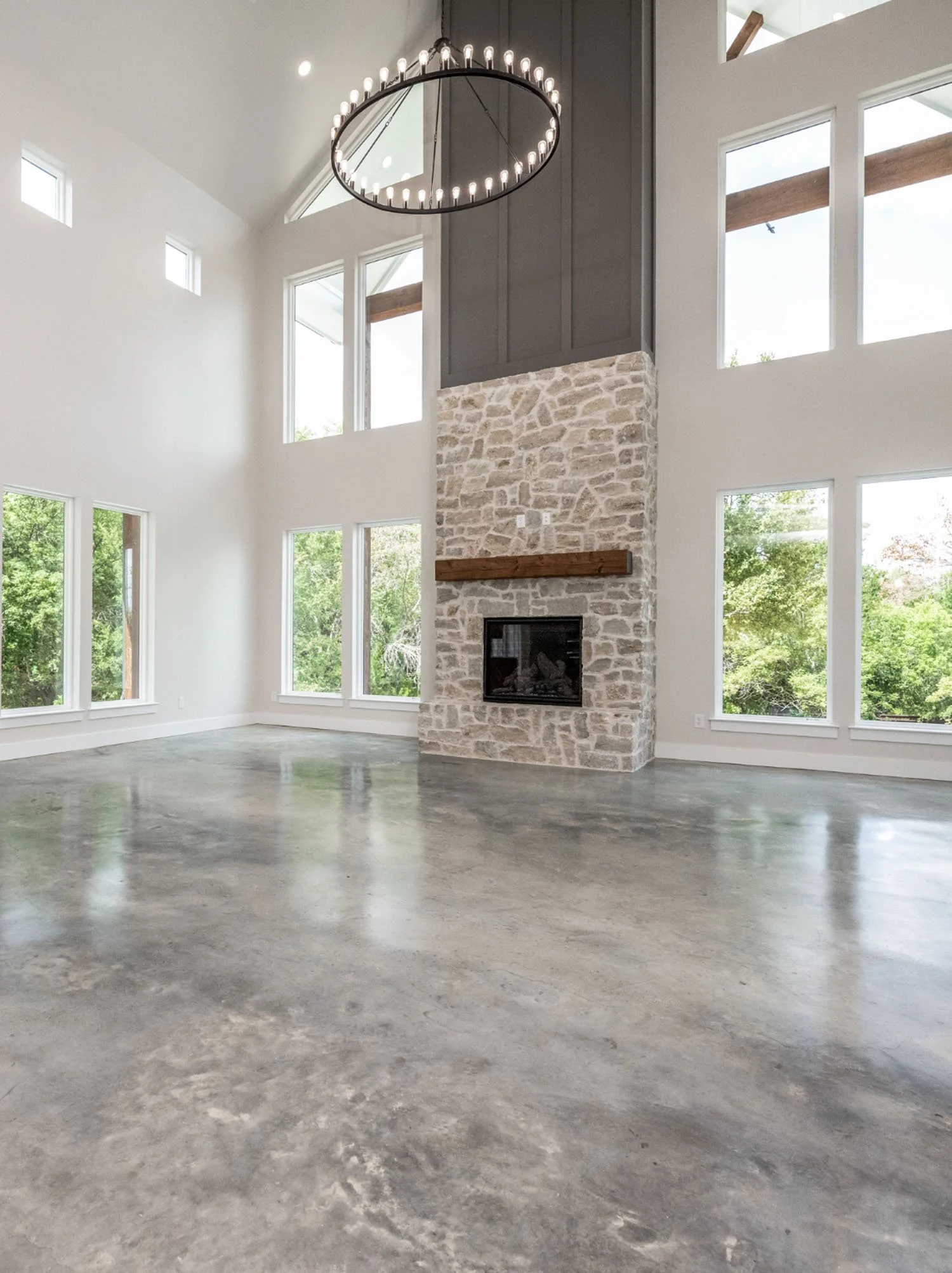 A spacious living room with polished concrete floors, a double-height vaulted ceiling, a large stone fireplace, and floor-to-ceiling windows.