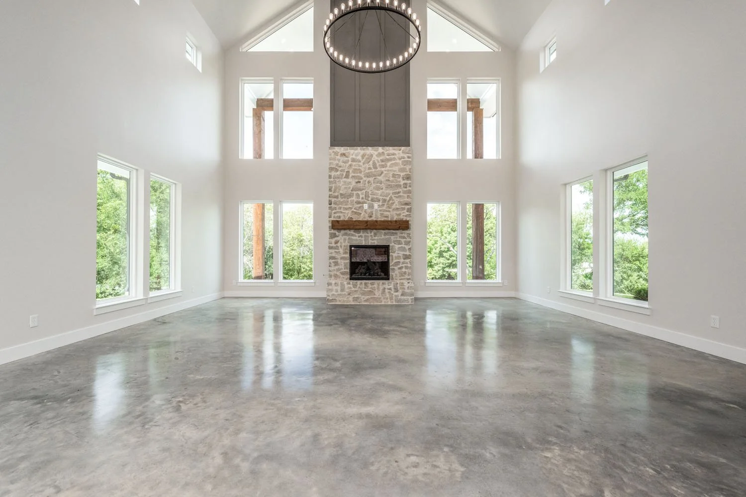 High-ceilinged living area featuring a stone fireplace, large windows, polished concrete flooring, and a round industrial-style chandelier.