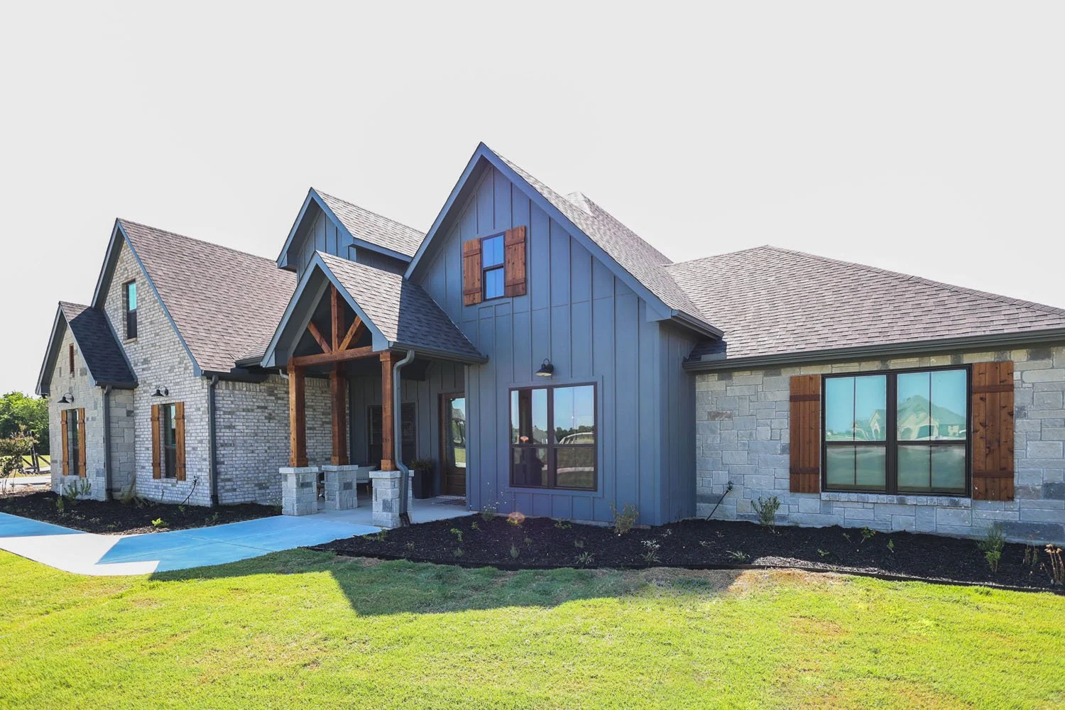 An angled view of the front exterior of a home featuring a mix of light stone and grey board-and-batten siding, rustic wooden shutters, and a covered entryway.