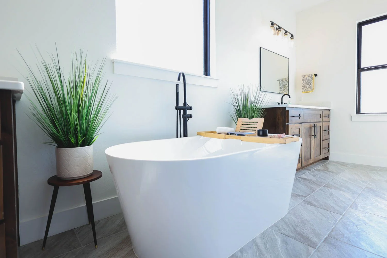 A modern bathroom featuring a white freestanding soaking tub with a bamboo tray, a dark wood vanity, and a large window overlooking green foliage.