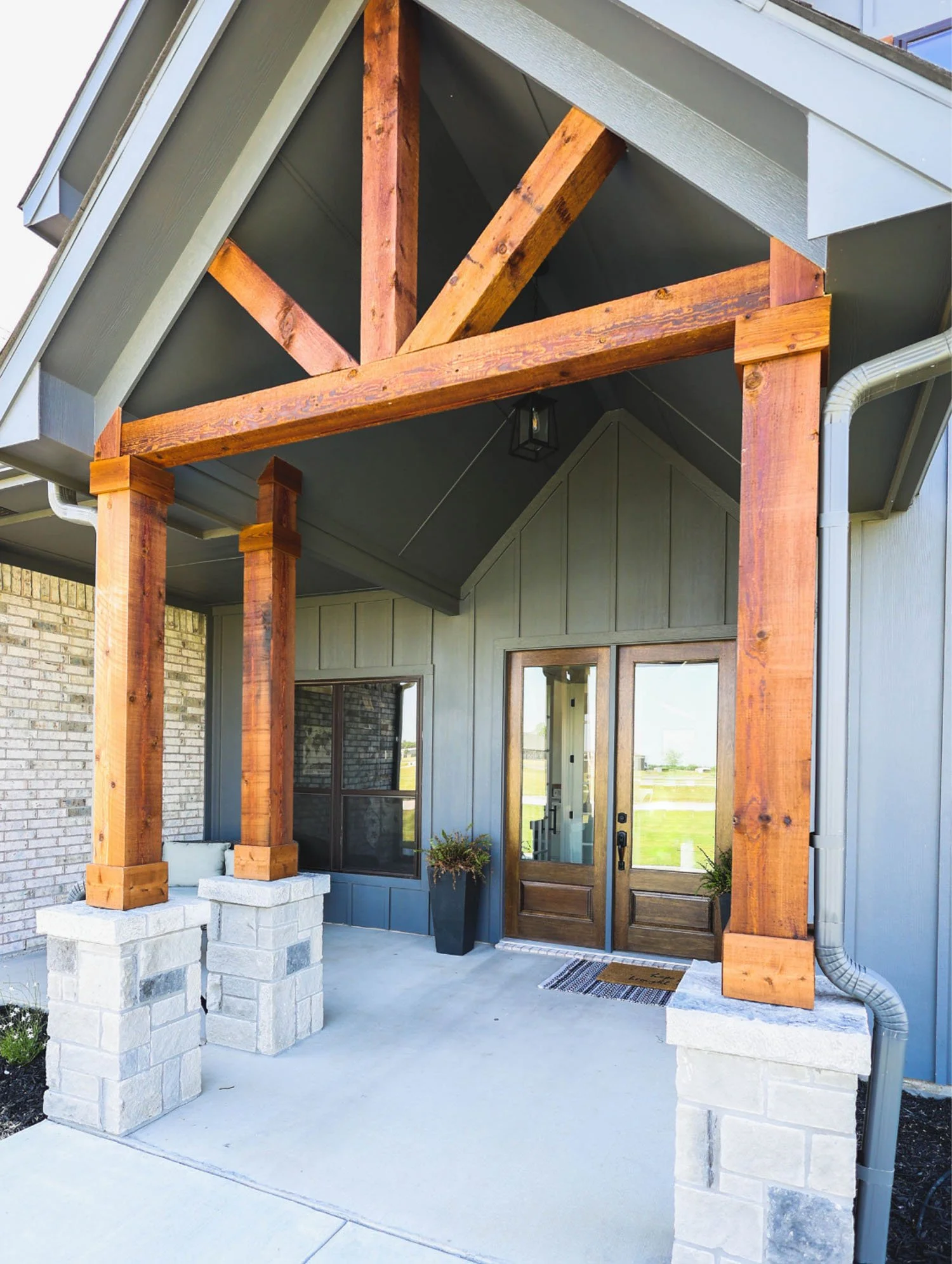 The front porch of a home featuring a dark gray exterior, large wood timber posts supported by stone bases, and a wooden double front door.