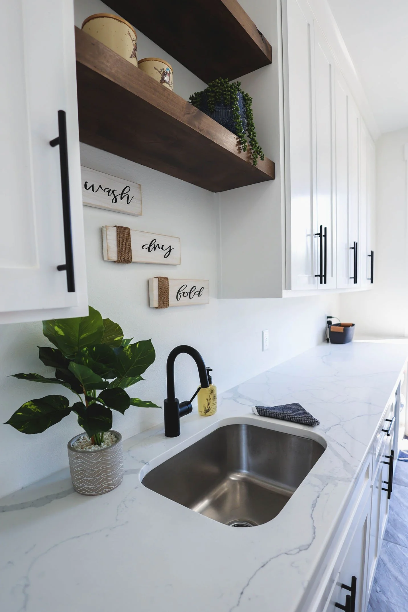 A bright laundry room featuring white cabinetry, floating wood shelves, a marble-look countertop, and a stainless steel sink with a black faucet.