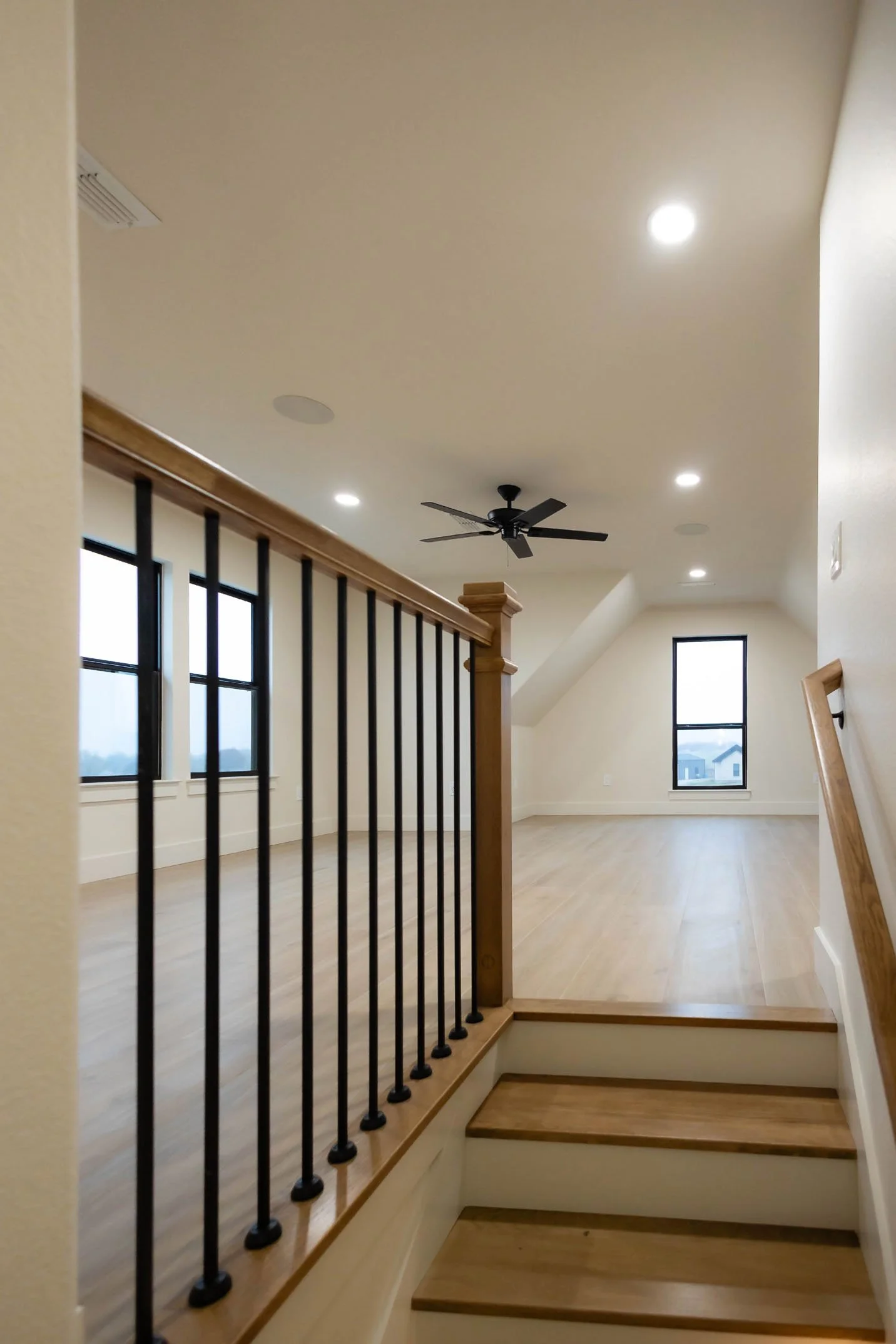 An open loft area with light wood flooring, a black ceiling fan, and a wood-and-black metal staircase railing, overlooking a bright room with large windows.