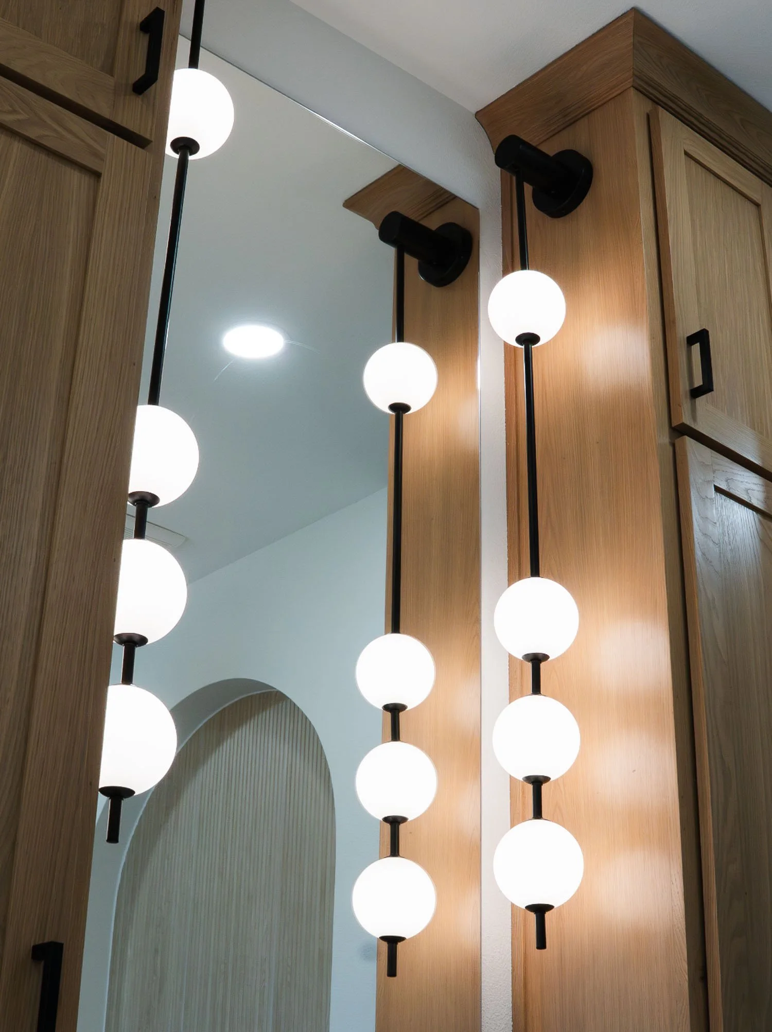 A close-up view of modern bathroom lighting fixtures with vertical stems and globe-shaped bulbs, mounted on light oak wood cabinets next to a mirror.
