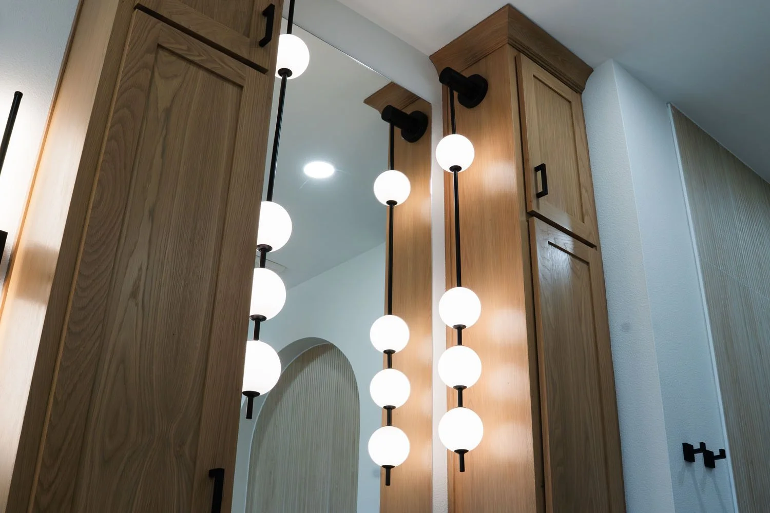 A side angle modern bathroom lighting fixtures with vertical stems and globe-shaped bulbs, mounted on light oak wood cabinets next to a mirror.