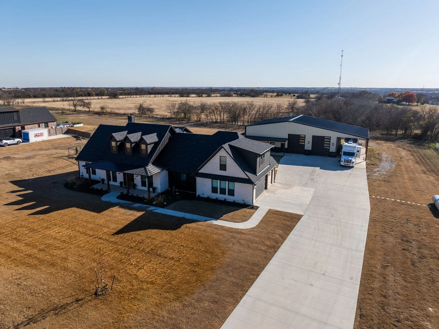 An aerial view of a custom home with a white brick exterior, black roof, and an adjacent large detached garage on a spacious rural lot.