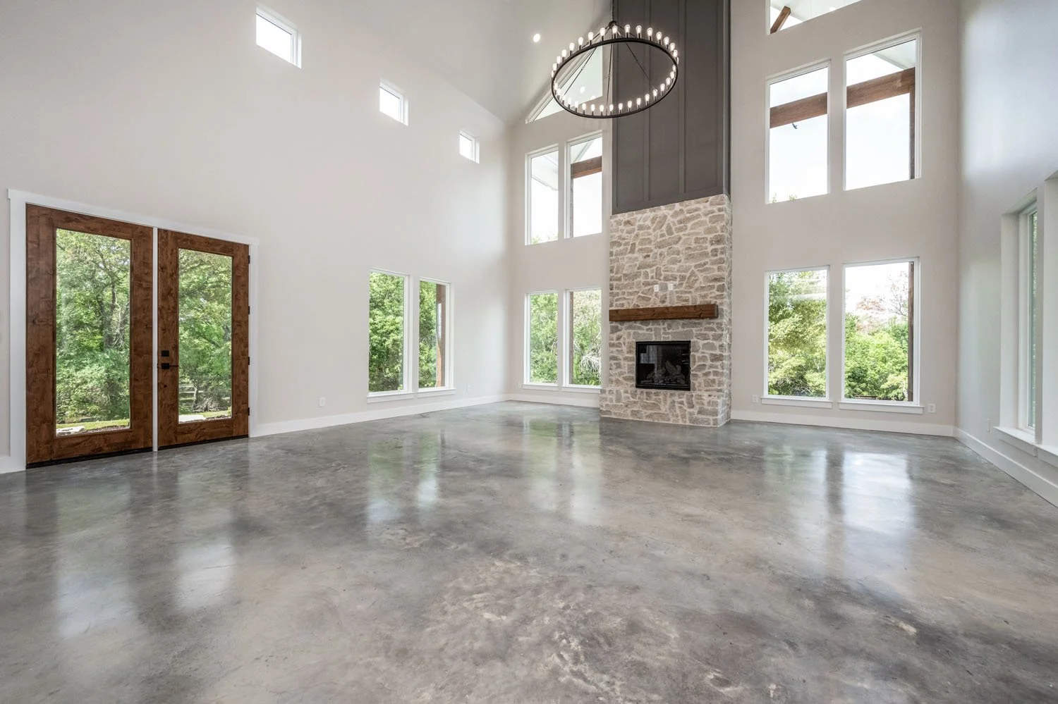 Large, vaulted living room with a prominent stone fireplace, dark grey accent wall, large windows, and a circular chandelier.