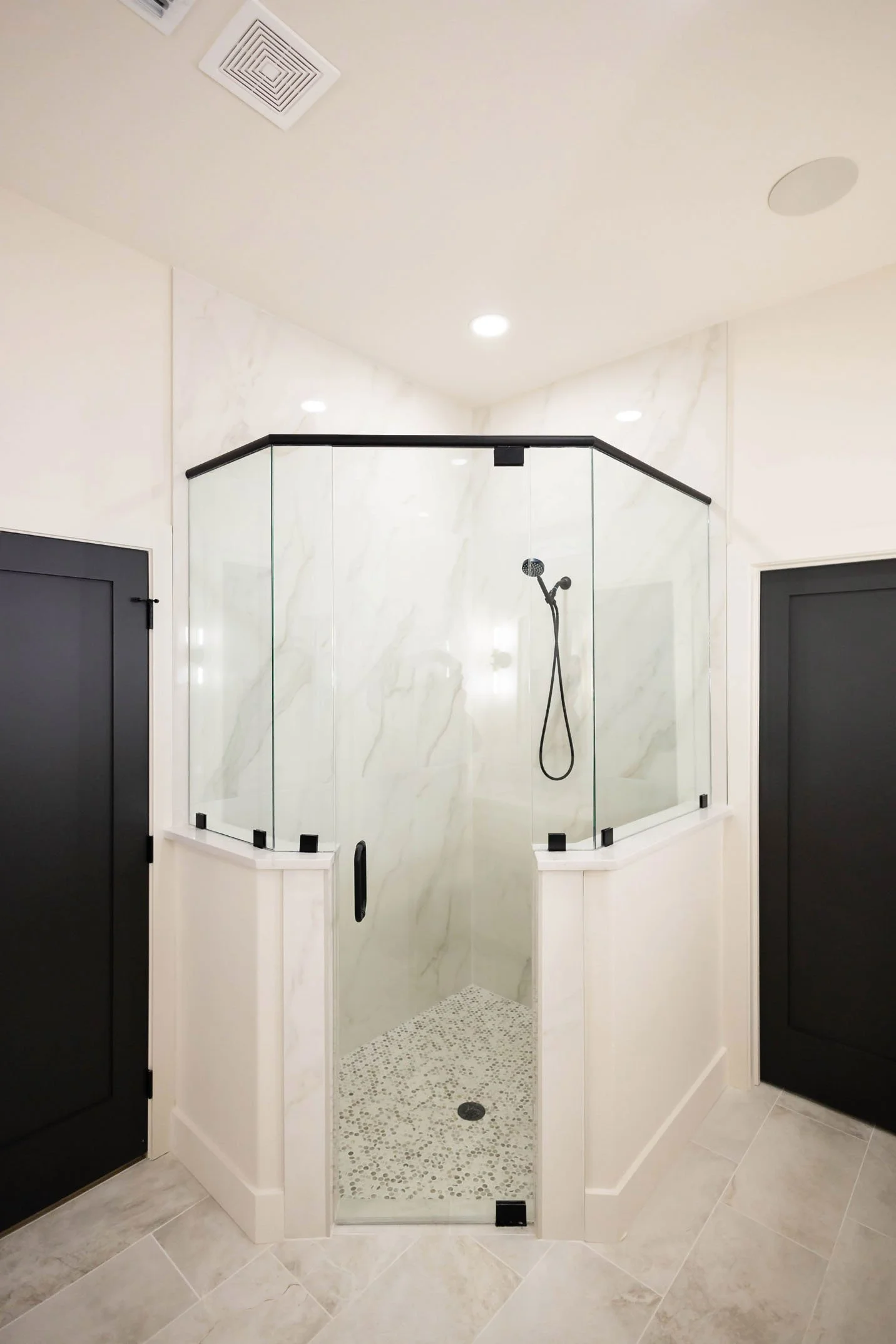 A corner shower featuring marble-look walls, a pebble-tiled floor, and a black glass enclosure, flanked by black doors.