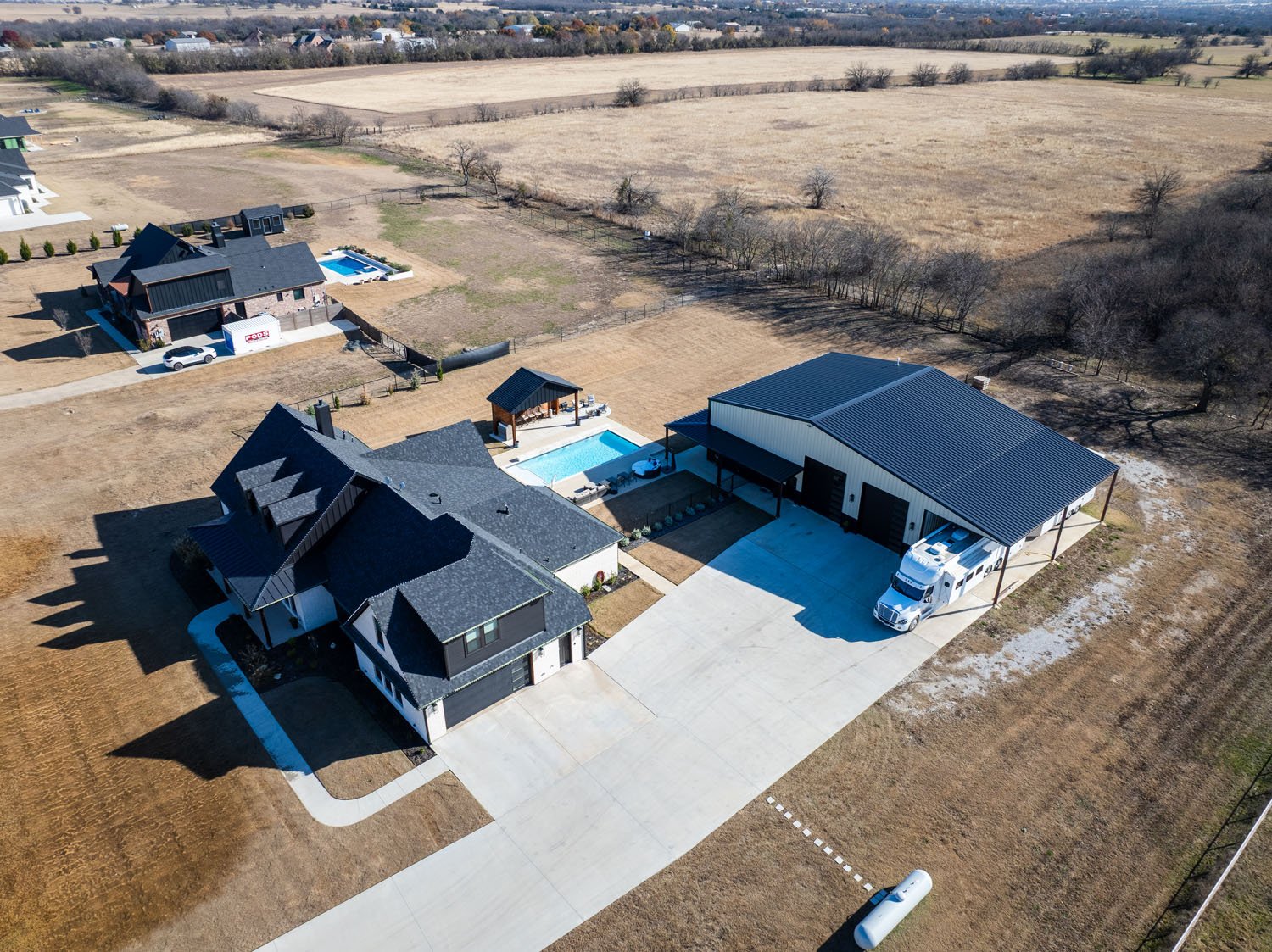 An elevated view of a custom house and large metal shop situated on a large rural plot of land with an open field.