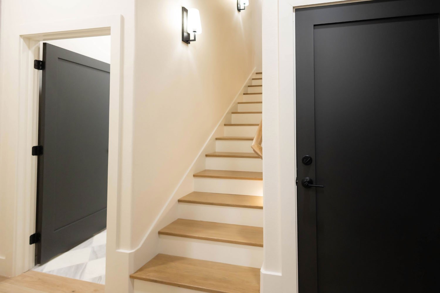 A staircase with light wood treads and white risers, situated between two matte black doors, with wall sconces providing warm lighting.