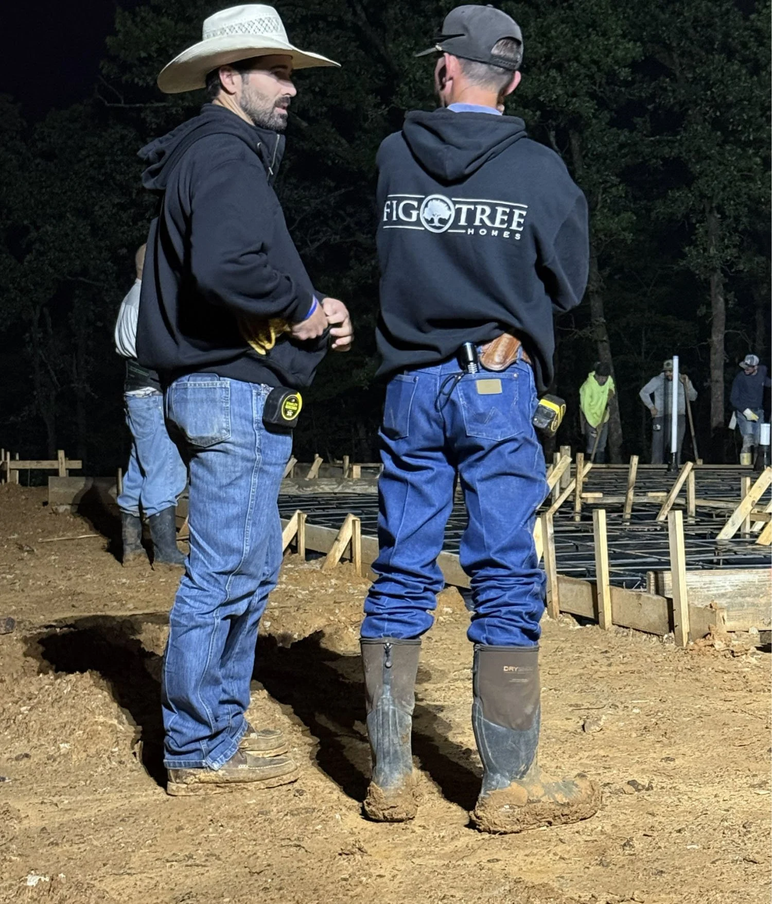 Two construction professionals, including founder Ian Sears, wearing Fig Tree Homes hoodies and work gear stand at a job site at night, overseeing foundation work.