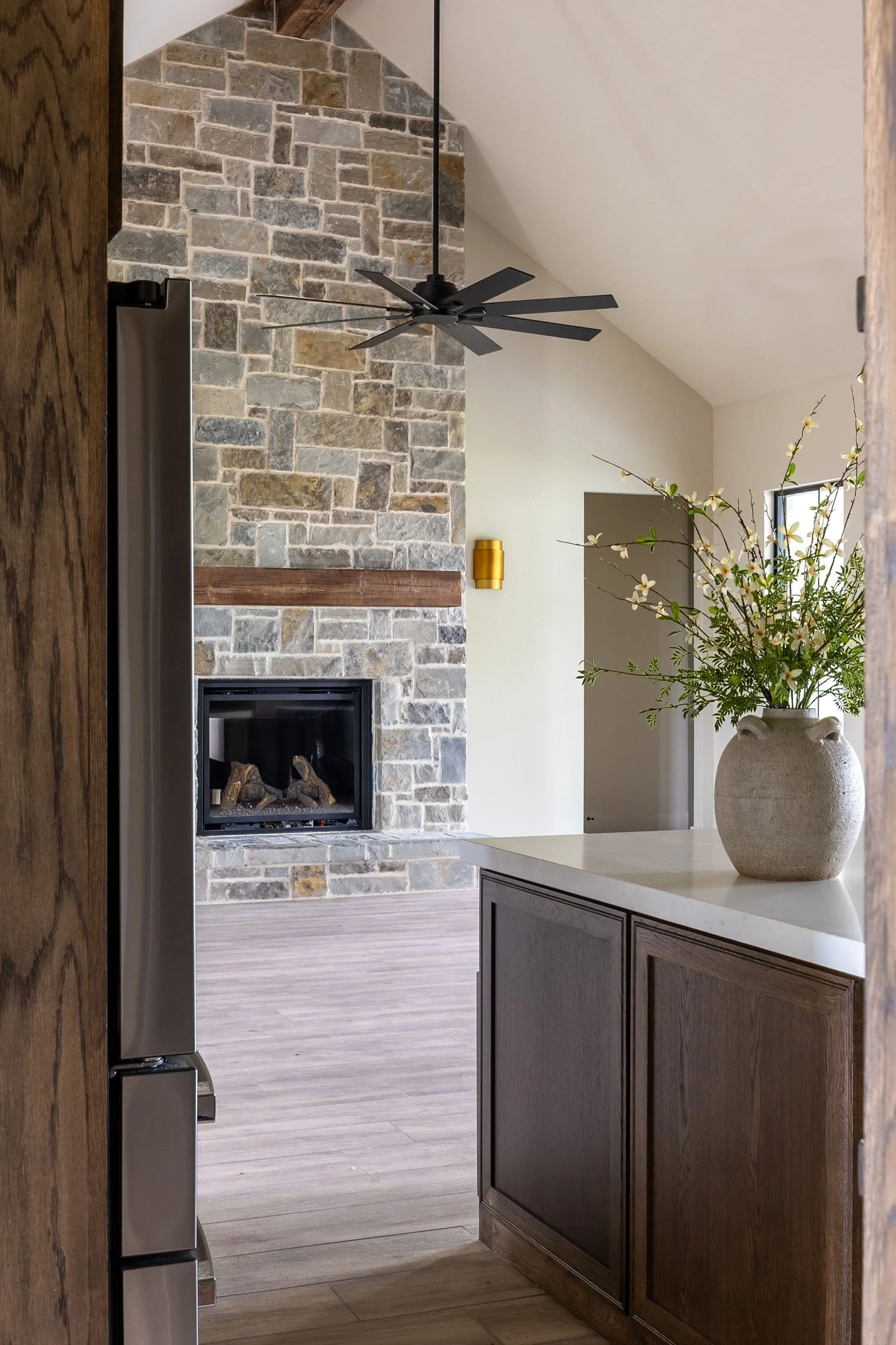 Modern living room view from a kitchen, featuring a floor-to-ceiling stone fireplace with a wooden mantel and a vaulted ceiling.