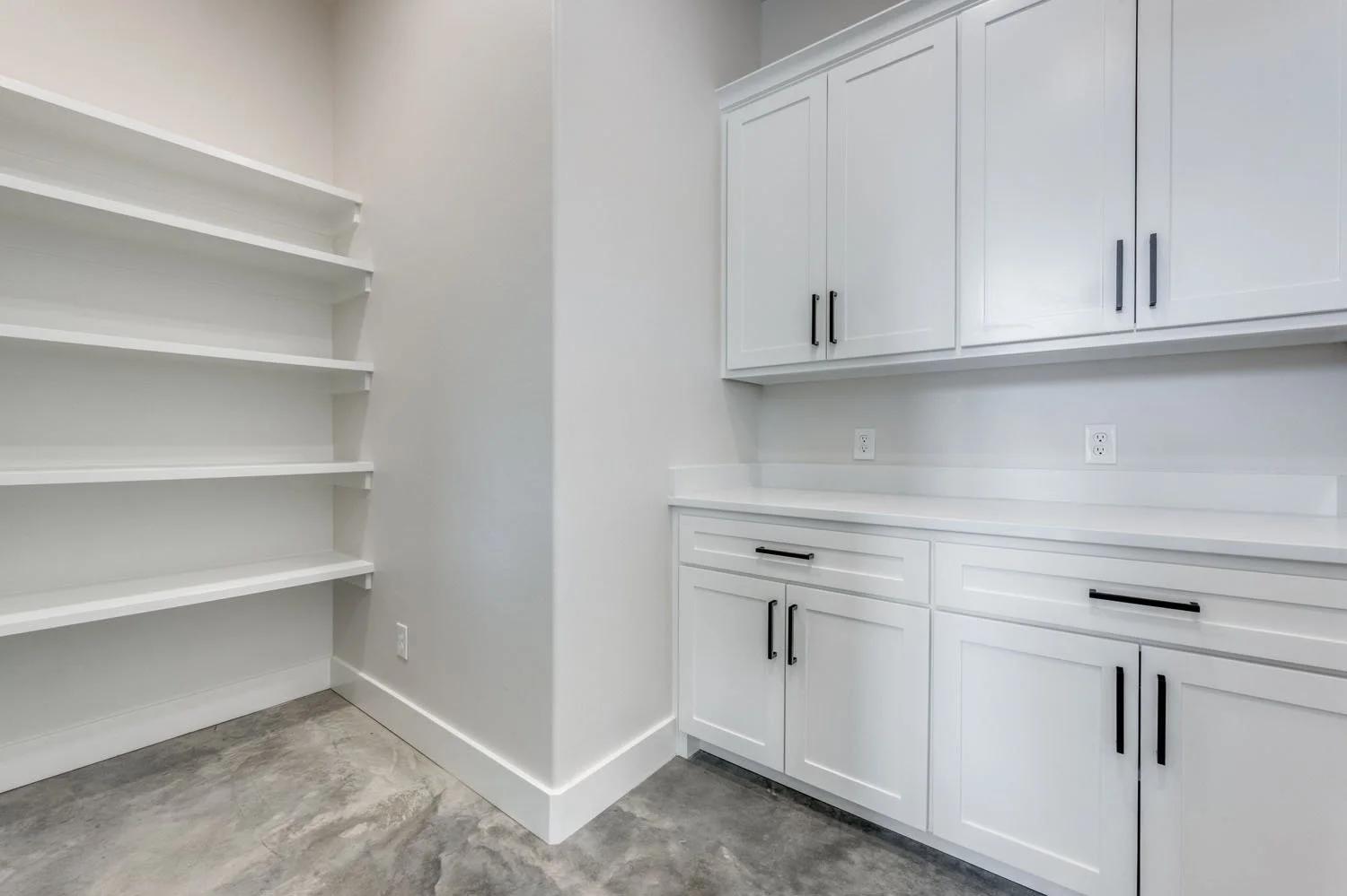 A clean, minimalist pantry room with white custom cabinetry, upper and lower storage, white countertops, and concrete flooring.