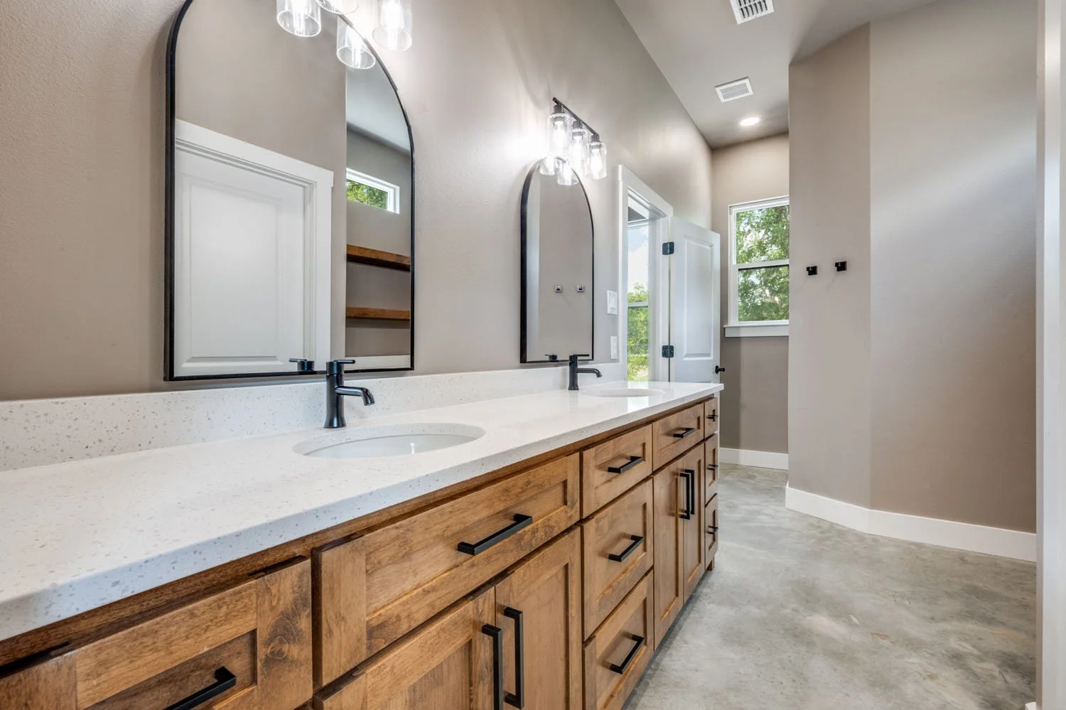 A bathroom vanity area with dual arched mirrors, black fixtures, a white quartz countertop, and warm-toned wood cabinetry against a neutral wall.