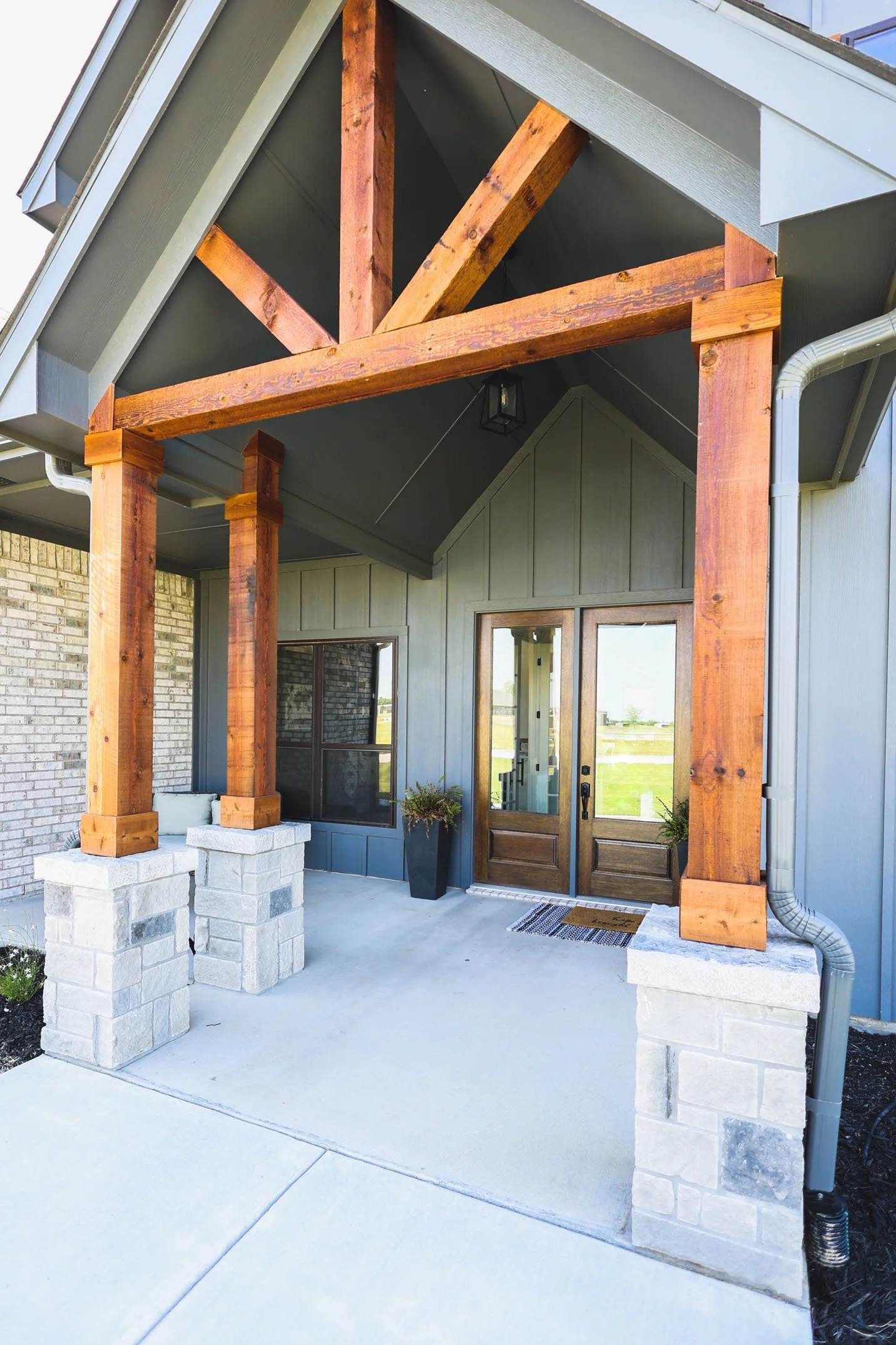The front entrance of a home featuring dark grey siding, a wooden front door, and a covered porch supported by rustic wooden beams over stone pillars.