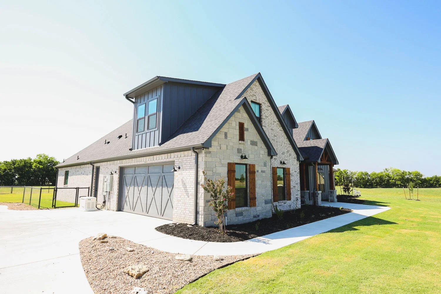A side view of a modern farmhouse-style home with light stone exterior, a grey garage, and dark metal roofing, set on a bright, sunny day.