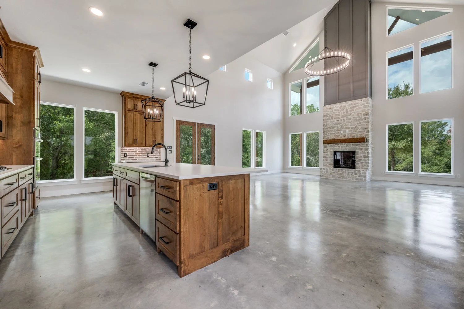 A spacious, open-concept kitchen and living area with a large wooden island, polished concrete floors, and a floor-to-ceiling stone fireplace.