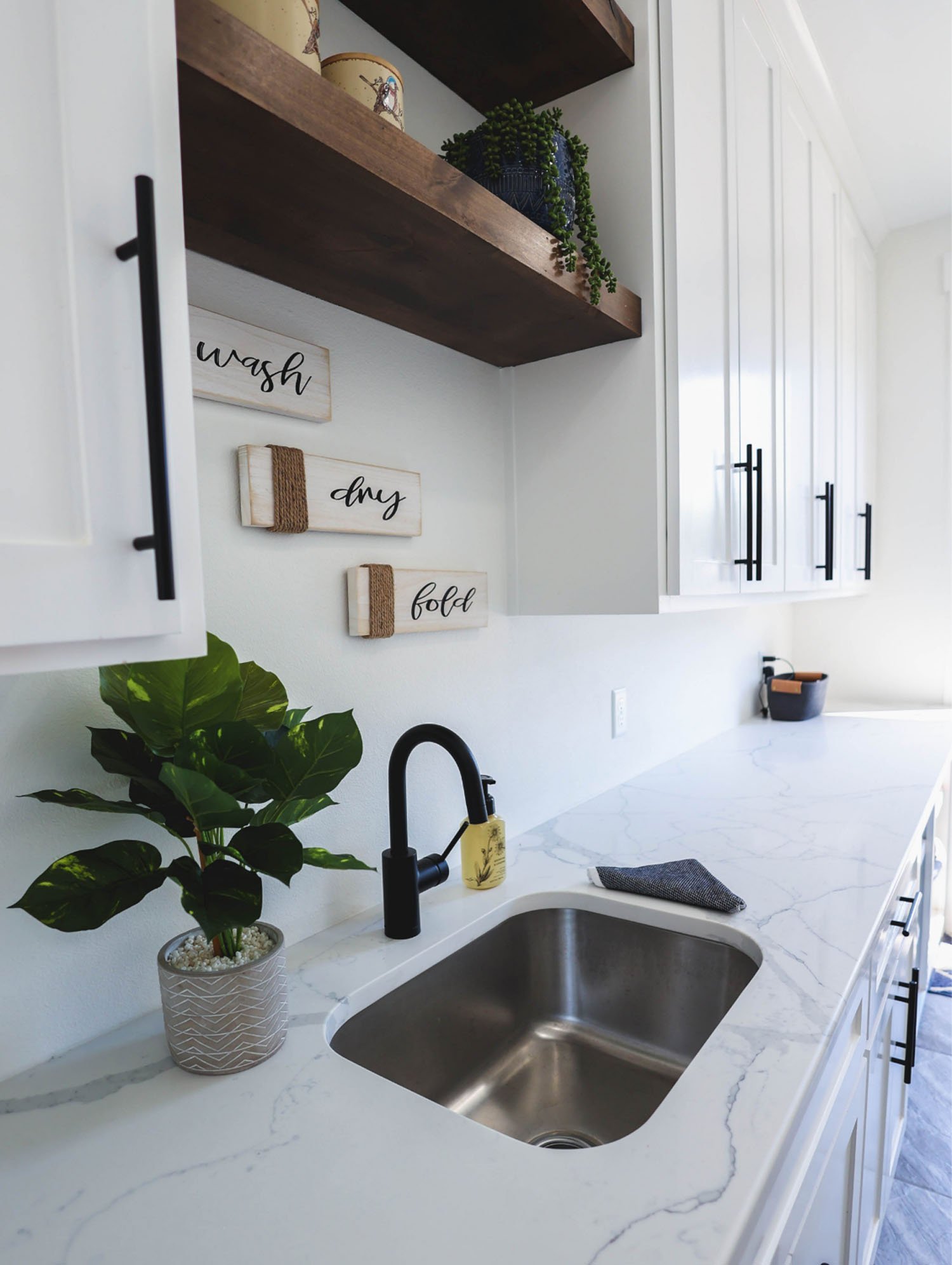 A bright laundry room featuring white cabinetry, marble countertops, a modern black faucet, and decorative 'wash, dry, fold' wall signage.