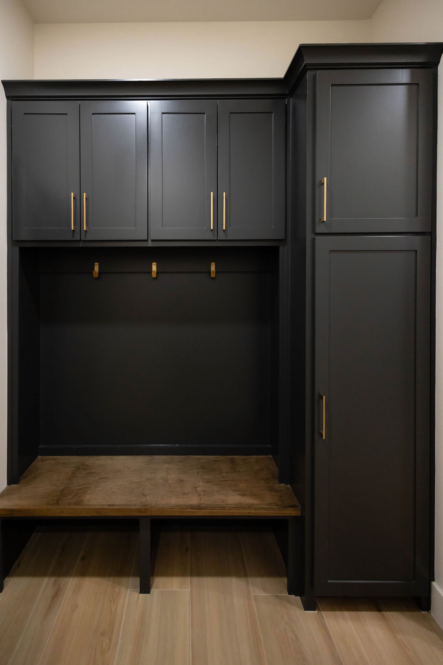 A mudroom locker unit featuring dark cabinetry, gold hooks, a wooden bench seat, and tall storage cabinets.