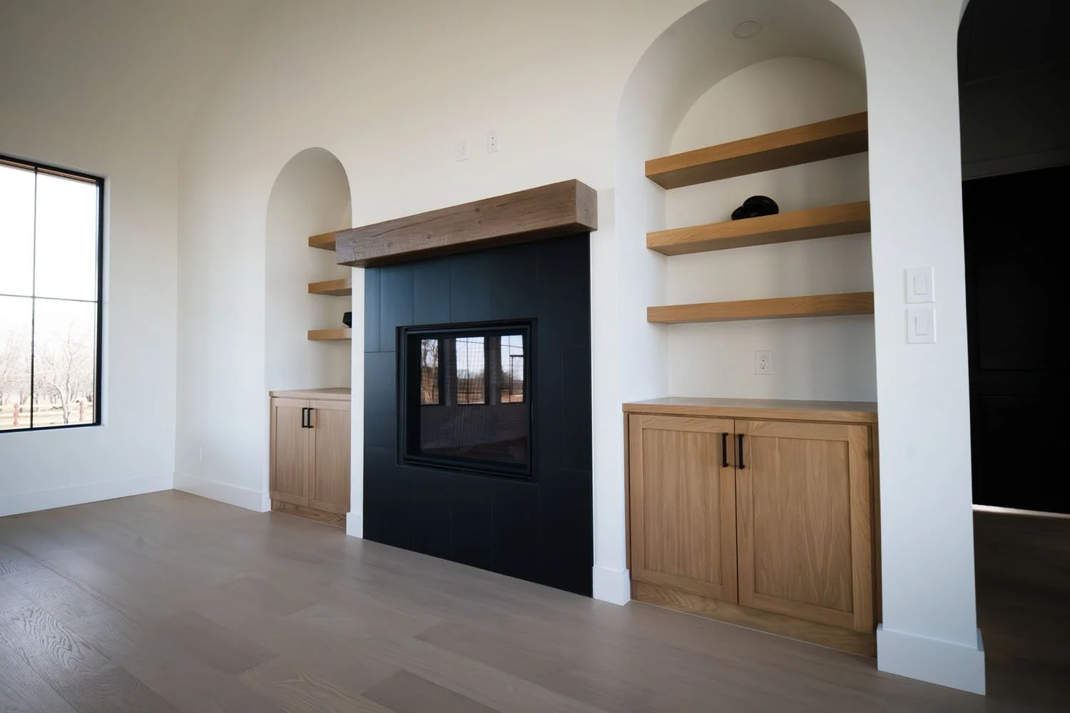 A bright living area featuring a centerpiece fireplace with a sleek black surround and a thick rustic wooden mantel, flanked by symmetrical arched built-in shelving and light wood cabinetry.