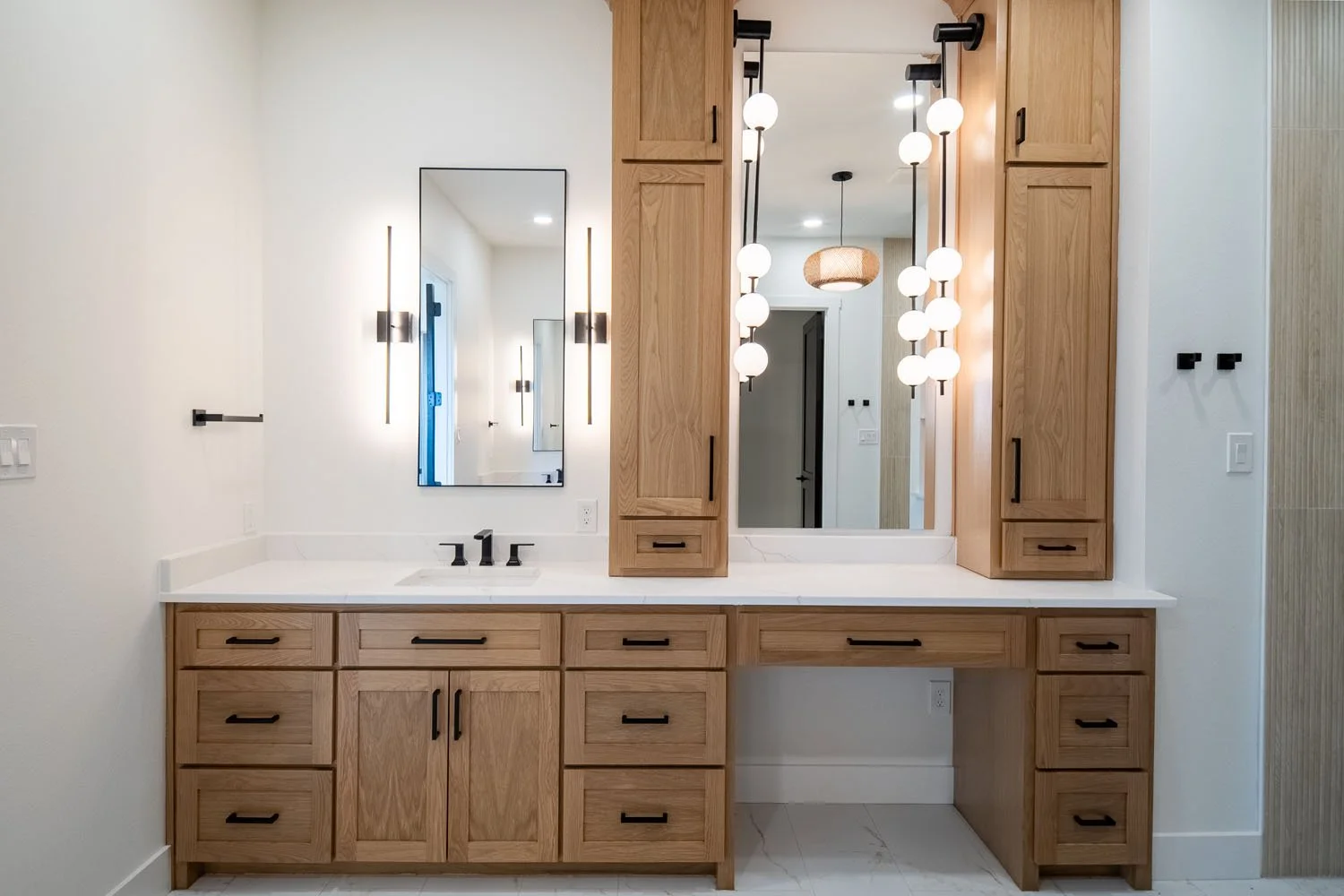 A bright, elegant primary bathroom vanity featuring natural oak cabinetry, white quartz countertops, dual tall linen cabinets, and contemporary multi-sphere vertical pendant lighting.