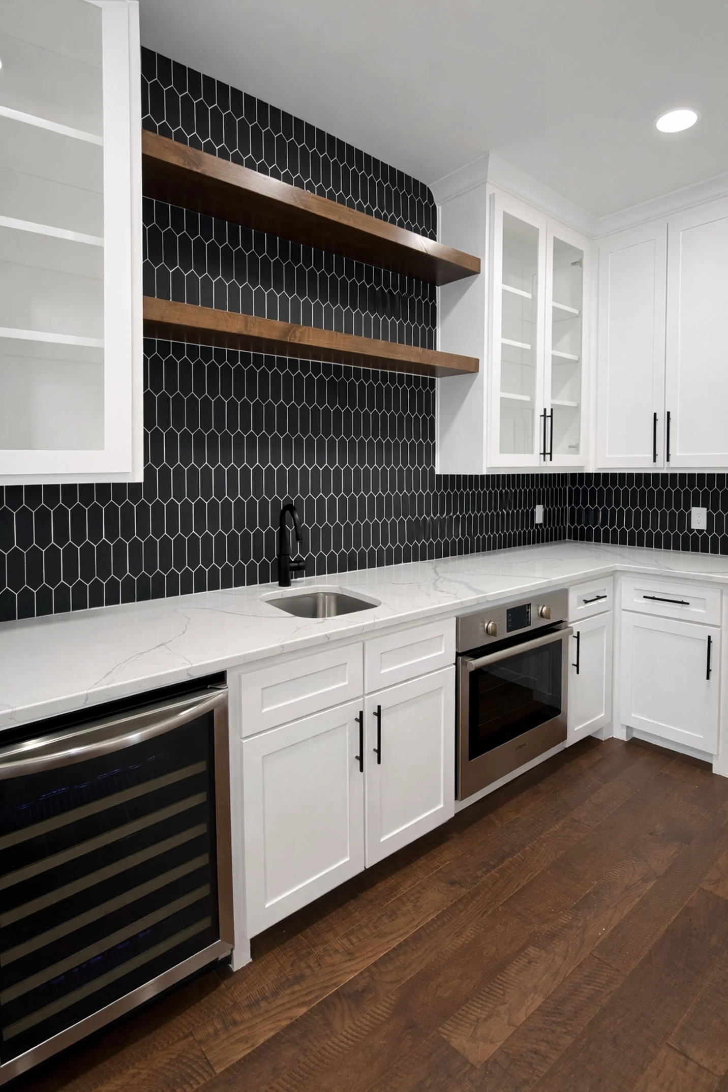 A modern kitchen space featuring white cabinets, a black geometric tile backsplash, floating wood shelves, and dark wood flooring.