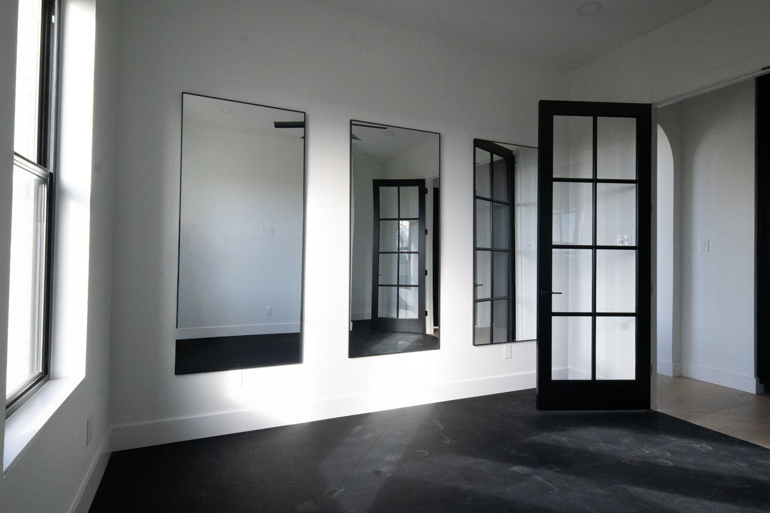A minimalist room interior with high-contrast design, featuring three rectangular wall mirrors, a black-framed glass paneled door, and dark flooring against crisp white walls.