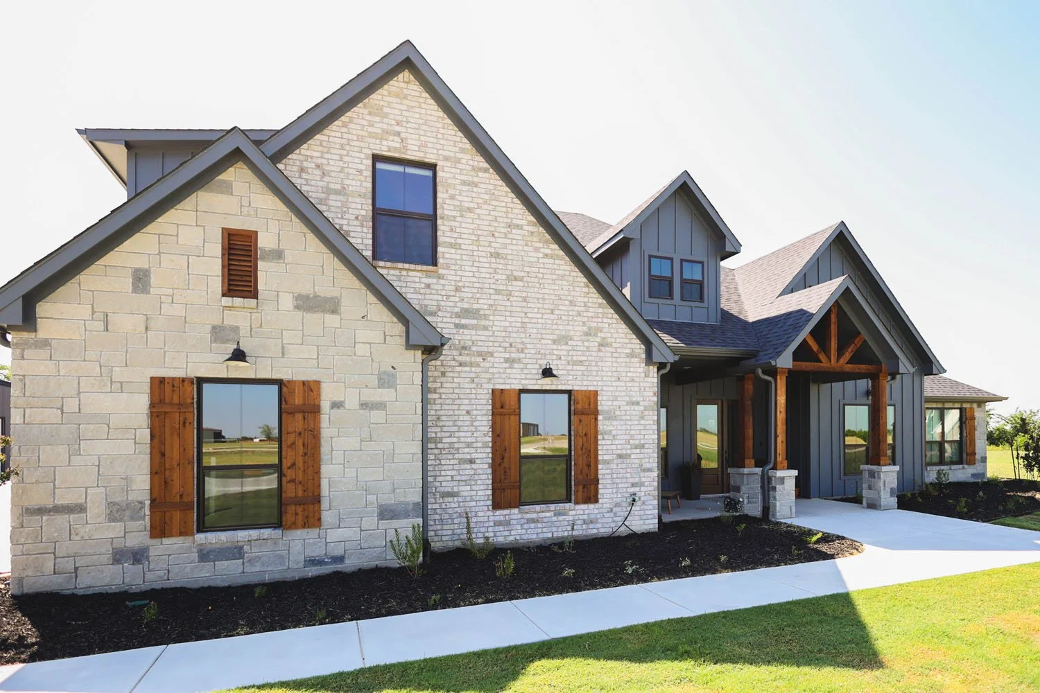 A modern farmhouse-style home exterior with a combination of light stone and dark grey siding, wooden shutters, and a covered front porch entryway.