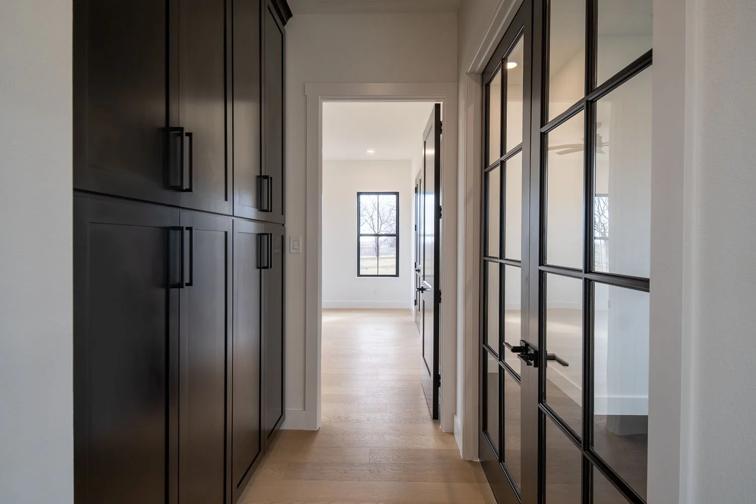 A bright interior hallway featuring floor-to-ceiling matte black custom cabinetry on one side and elegant black-framed glass double doors on the other, leading to a sunlit room.
